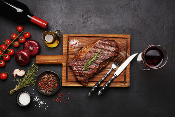 Obraz na płótnie Canvas Grilled Striploin beef steak on wooden board with bottle and glass of red wine, vegetables, herbs and spices over black concrete background. Top view