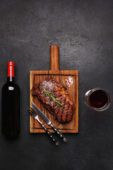 Grilled striploin beef steak on wooden board with bottle and glass of red wine, fork and knife over...