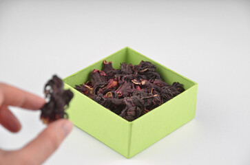 Egyptian hibiscus dry red tea (karkade) in a hand in a green box on a white background, closeup, macro, side view