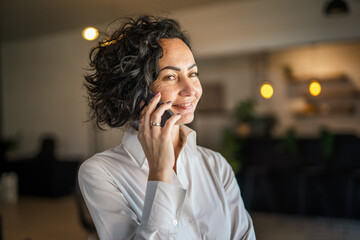 Fototapeta na wymiar One woman mature caucasian female businesswoman entrepreneur stand at work or home use mobile phone making a call talk real people copy space wear white shirt curly hair happy smile