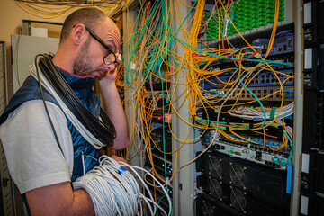 A technician technician with glasses is thinking about how to switch the Internet cables in a data center. A man with a large skein of wires is working in a server room.