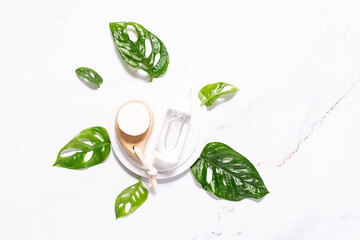 Skin care serum in white glass bottle with dropper and soft face brush on white marble background decorated by beautiful green monstera leaves.