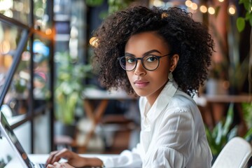 Focused African American girl working on laptop PC. Beautiful young woman wearing glasses and white blouse sitting at table looking at computer screen and writing in modern office