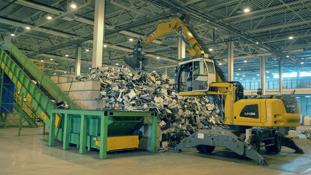 Yellow excavator puts trash onto conveyor at a factory. Recycle process, secondary use of plastic.