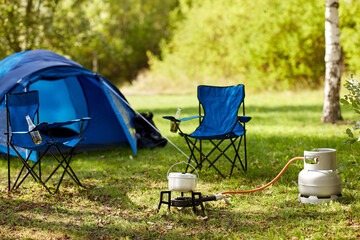 food cooking, tourism and travel concept - close up of camping pot on tourist gas burner, blue tent and two folding chairs at camp - 759995739