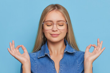 Golden-haired pretty girl in glasses and blue shirt standing isolated doing yoga mudra with closed eyes and smile on face, meditation concept, copy space