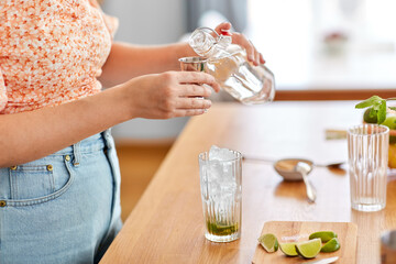 drinks and people concept - close up of woman pouring water from glass bottle to jigger and making lime mojito cocktail at home kitchen