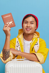 Happy Asian guy shows his passport posing with his travel suitcase dressed in yellow raincoat and red hat, travel time concept, copy space