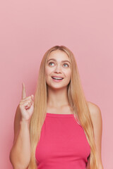 Beautiful girl model against pink background, long golden hair, blue eyes, finger pointing upwards with cute smile, happy life concept, copy space