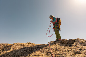 A woman is standing on a rocky hill, looking at a rope. She is wearing a backpack and a helmet. The scene is peaceful and serene, with the woman focused on her task