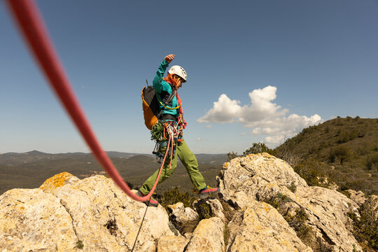 A woman is climbing a mountain with a red rope attached to him. She is wearing a backpack and a helmet. The scene is adventurous and exciting, as the woman is taking on a challenging climb