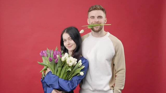 Shocked lady and positive man holding white fresh tulips, looking at camera isolated on blue background