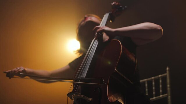 Crop of emotional female musician playing cello with bow while sitting on stage in bright light. Adorable woman performing with musical show and expressing deep feeling while enjoying music
