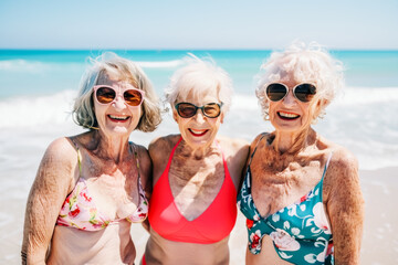 Three elderly women with sunglasses and smiling are standing side by side on a sandy beach. Retirement and summer concepts.
