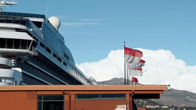 a huge cruise ship stands at the pier, mountains in the background, Flags of principality of Monaco in the wind at sunny weather, the port of Hercules