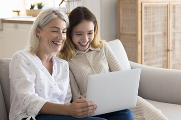 Cheerful mature mother spend time with teen daughter, sit on couch with laptop, watch funny video...