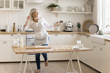 Cheerful mature woman in apron dancing in the kitchen, listen favorite music, moving, feel carefree, prepare holiday cake for family, enjoy culinary hobby alone at home. Domestic activity, cooking - obrazy, fototapety, plakaty