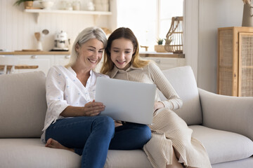 Carefree pre-teen girl spend time with 45s mother at home, sit on sofa with wireless computer,...