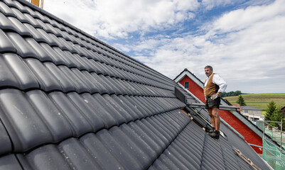 roofer in traditional German clothing of a craftsman working with roof tiles on the roof. - 760056769