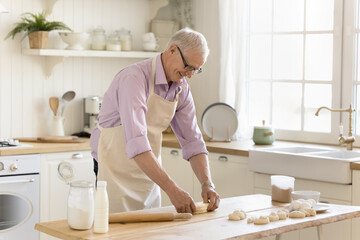 Smiling old single man in eyeglasses wear apron cooking alone in cozy domestic kitchen prepare dough for homemade pastry, makes handmade dessert on weekend at home. Culinary, recipe, natural products