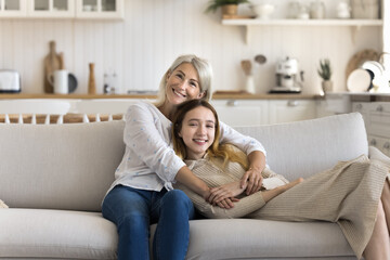 Mature 45s mother cuddling pre-teen 14s daughter resting in living room, smile, looking at camera...
