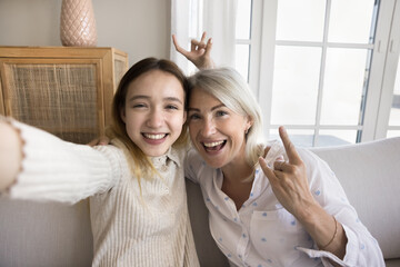 Middle-aged mum and pre-teen daughter hold device, smile, show hand gestures looking at camera,...
