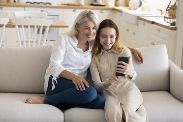 Smiling teen girl rest at home with mature mother, hold mobile phone, chatting online, spend time in social media, browse internet, watch funny videos. Family having pleasant weekend use modern apps