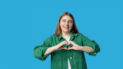 Attractive 30s young woman feels happy and romantic shapes heart gesture expresses tender feelings poses against blue background. Emotional face. Human emotions, facial expression concept