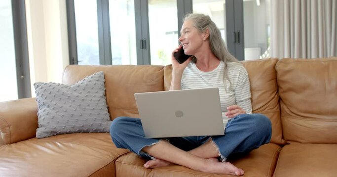 A mature Caucasian woman chats on her phone, laptop on her lap, on a couch at home