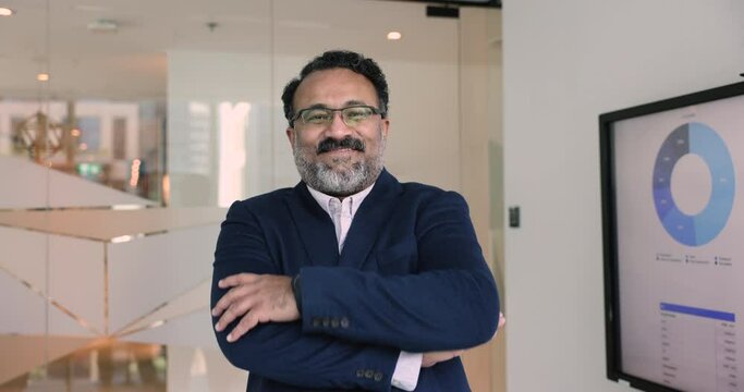 Mature Indian businessman in formal suit posing in office with arms-crossed, smile looking at camera, exudes confidence and leadership, portrays reliability and expertise in finances and accounting