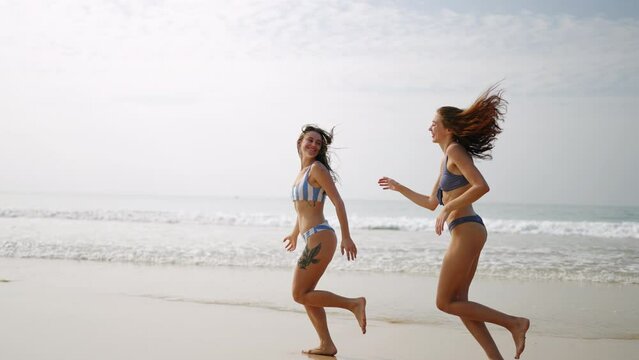 Two happy girls in bikini running at sea water waves. Cheerful female friends having fun, tanning on summer day at seaside. Pretty young models run at the ocean. Girls in swimwear run in slow motion.
