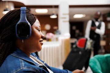 Smiling African American woman wearing wireless headphones resting in hotel lobby. Happy female tourist listening to music while waiting for check-in at resort, arriving on vacation