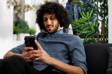 Company project manager writing message to coworkers using smartphone in business office. Start up young arab entrepreneur relaxing and chatting in social media on smartphone - obrazy, fototapety, plakaty