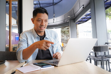 Asian man happy and smile face with blue shirt using laptop and mobile phone in coffee shop, online freelance business