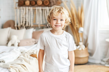 Caucasian 5 year old boy in white t-shirt standing next to his bed in a room with Nordic style decor. Copy space.