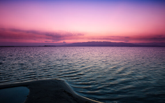 A boat's bow gently cuts through the waters of Delta del Ebro under a spectacular sunset sky painted in shades of pink and purple