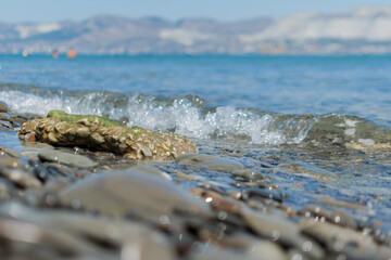 Seascape - wet round pebbles in the foreground are covered with a wave of the blue sea, mountain...
