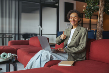 a woman is sitting on a red couch with a laptop and giving a thumbs up in nice modern coworking office