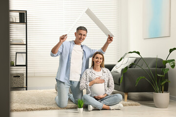 Young family housing concept. Man holding cardboard roof over his pregnant wife head at home