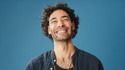 Close up of laughing attractive man with curly hair, dressed in blue shirt, crossing his arms and looking at camera, isolated on blue background in studio