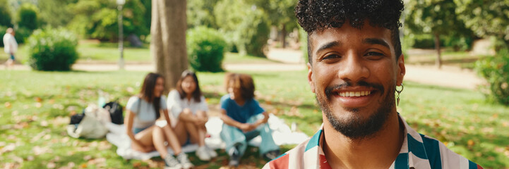 Close-up of young man with curly hair wearing striped shirt sitting in park having picnic on summer day outdoors, smiling and talking with friends, Panorama