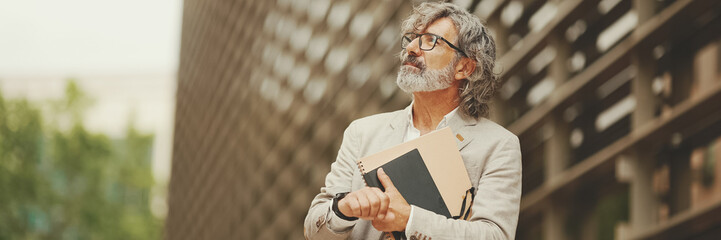 Panorama of mature businessman holding folder with business documents is standing outside on modern...