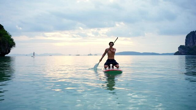 4K Young Asian man swimming and rowing paddle board in the ocean at tropical island at sunset. People enjoy outdoor active lifestyle water sport surfing and paddle boarding on summer beach vacation.