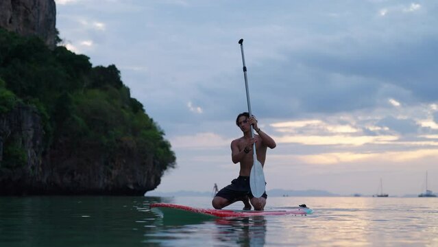 4K Young Asian man swimming and rowing paddle board in the ocean at tropical island at sunset. People enjoy outdoor active lifestyle water sport surfing and paddle boarding on summer beach vacation.