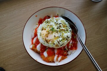 A bowl filled with rice, chicken katdu and eggs on the top. Served on wooden table. Sauce and mayonaise on the top.