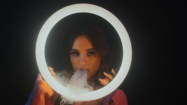 Medium portrait of gen Z girl smoking vape and looking at camera behind ring light