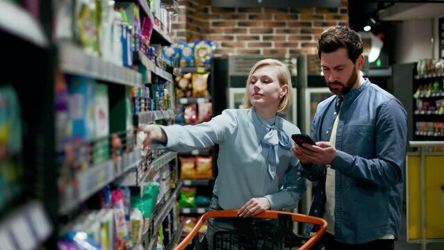 Front view of friendly caucasian marriage people doing shopping with list on portable smartphone indoors. Attractive wife taking package on napkins in aisle of household chemicals. Concept of retail.