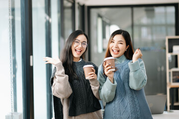 Happy two Asian business woman holding coffee cup in coworking office