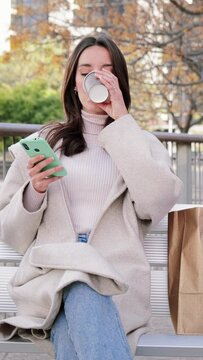 Vertical. Happy young woman sitting on a bech using a smartphone to text messages after shopping on-line. female looking to the cellfone device sharing content on social media and drinking coffee