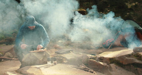 A man cooks food outdoors on a camping trip while sitting by a campfire. Cooking food on a tourist trip to the mountains. Camping and outdoor cooking.